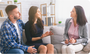 A couple sits on a couch talking to a professional woman with a notepad, suggesting an opioid addiction treatment or counseling session.