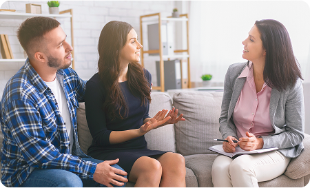 A couple sits on a couch talking to a professional woman with a notepad, suggesting an opioid addiction treatment or counseling session.