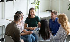 A group of six people sits in a circle having a discussion about opioid addiction treatment in a bright room with shelves and a potted plant in the background.