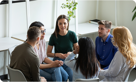 A group of six people sits in a circle having a discussion about opioid addiction treatment in a bright room with shelves and a potted plant in the background.