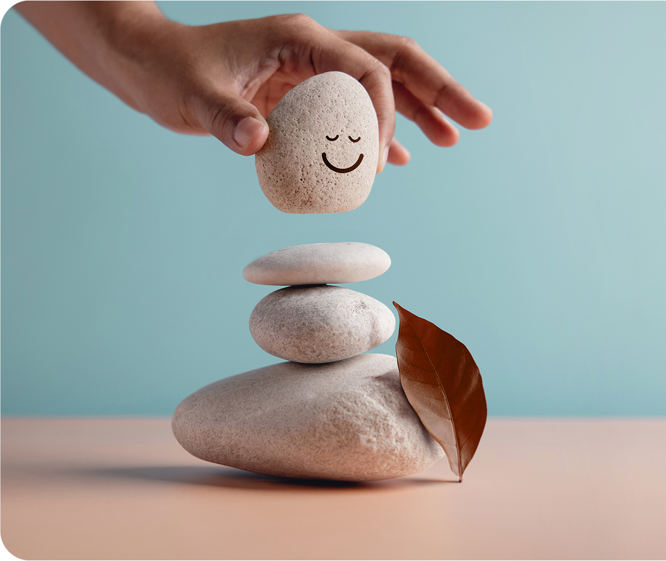 A hand places a stone with a smiling face drawn on it atop a stack of three smooth stones, with a brown leaf resting on them—symbolizing hope and balance found through Suboxone Addiction Treatment.