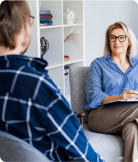 A woman with glasses sits in a chair holding a clipboard, listening to a man discuss opioid addiction treatment during their conversation in a modern office setting.