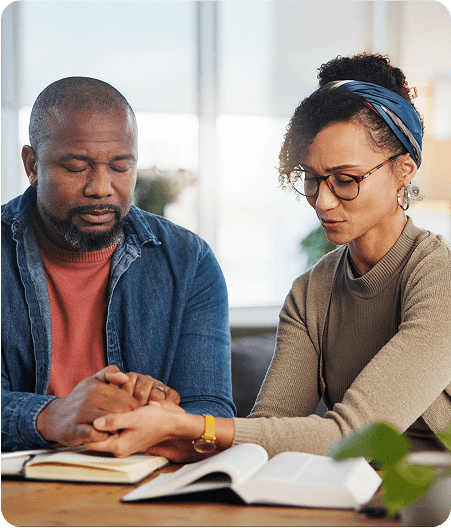 Two people sit at a table holding hands with closed eyes, appearing to pray, with an open book and notebook in front of them—reflecting on their journey through opioid addiction treatment and healing together.