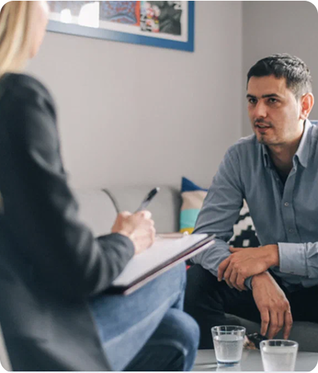 A man sits on a couch talking to a woman who is taking notes on a clipboard; two glasses of water are on the table between them, suggesting a discussion about Suboxone Addiction Treatment or other medically assisted treatment options.