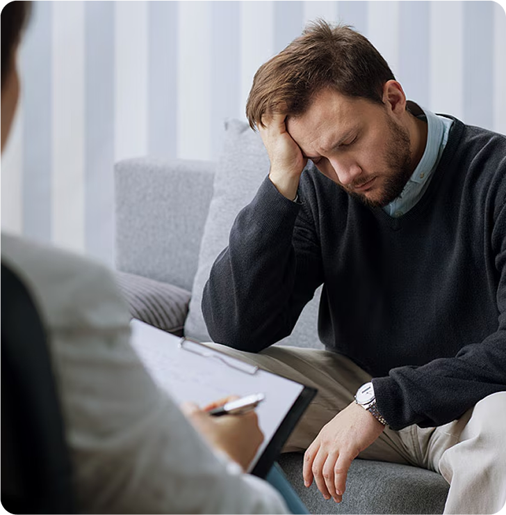 A man sits on a couch with his head resting on his hand, appearing distressed, while another person takes notes on a clipboard in the foreground during Suboxone Addiction Treatment.