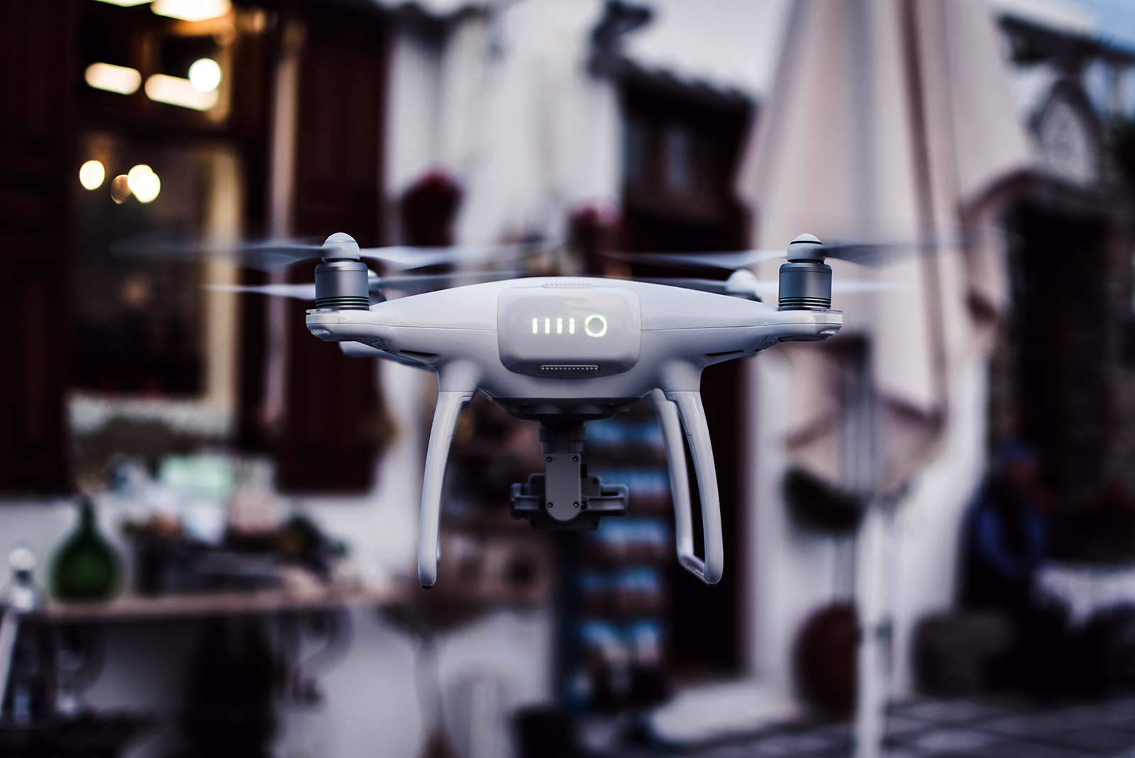 A white quadcopter drone with a camera hovers outdoors, its steady flight as precise as Medically Assisted Treatment for opioid addiction, with buildings and a blurred background visible.